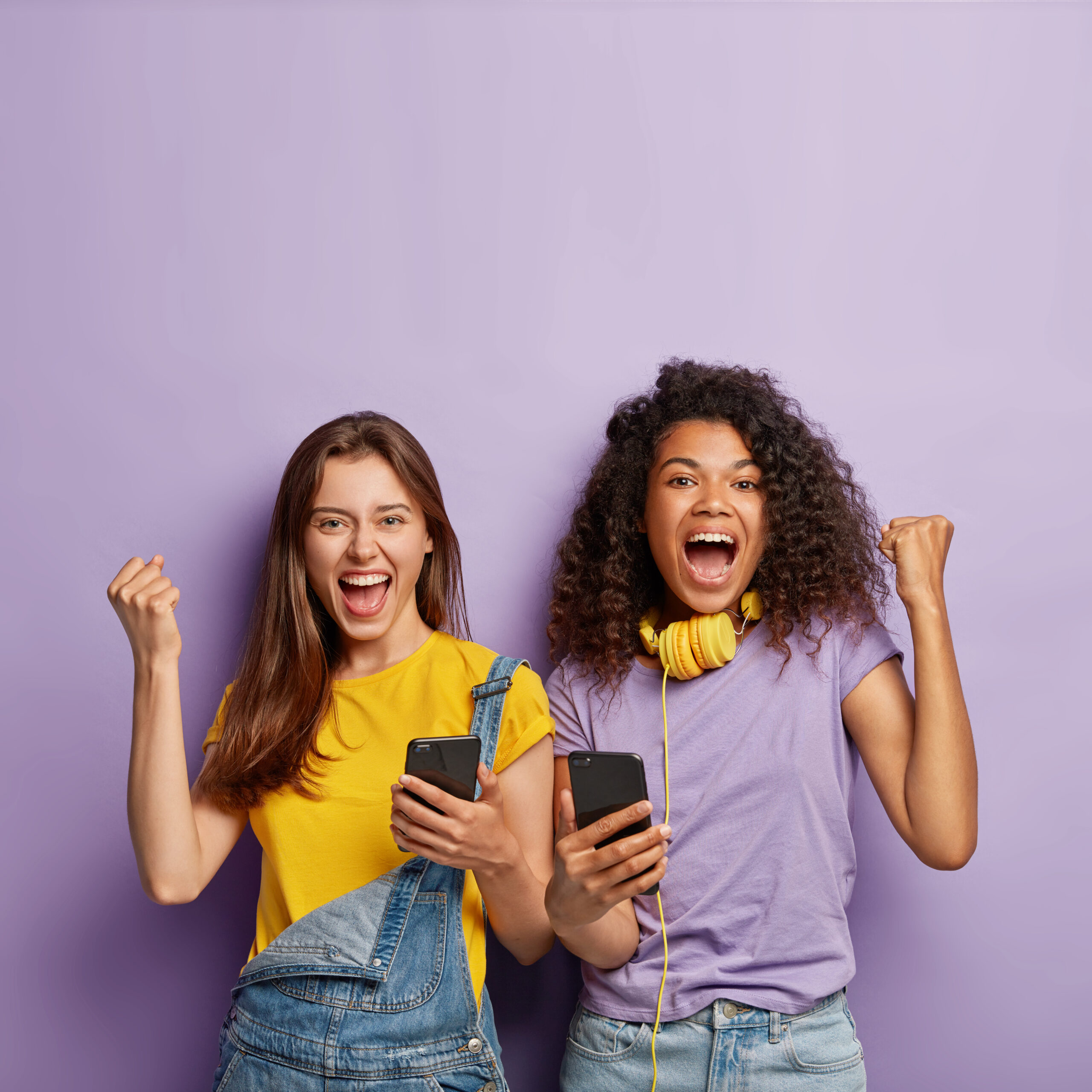Optimistic mixed race female classmates clench fists from joy, receive confirmation of entering university, show success gesture, hold smartphones, isolated on purple wall, smile with opened mouthes
